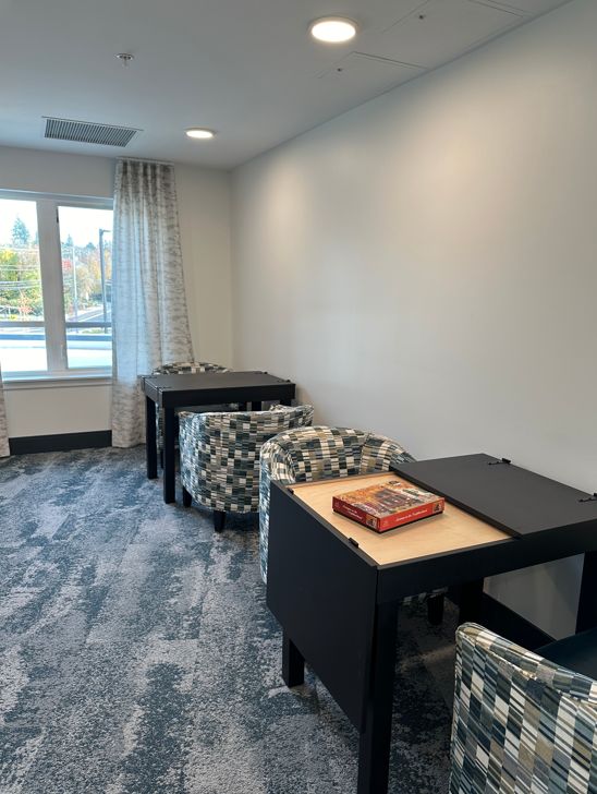 A modern community room setup with 2 black rectangular jigsaw puzzle tables, patterned armchairs, and a blue-gray carpeted floor. A window with white curtains is visible on the left, and a red puzzle box sits on the tabletop in the foreground.