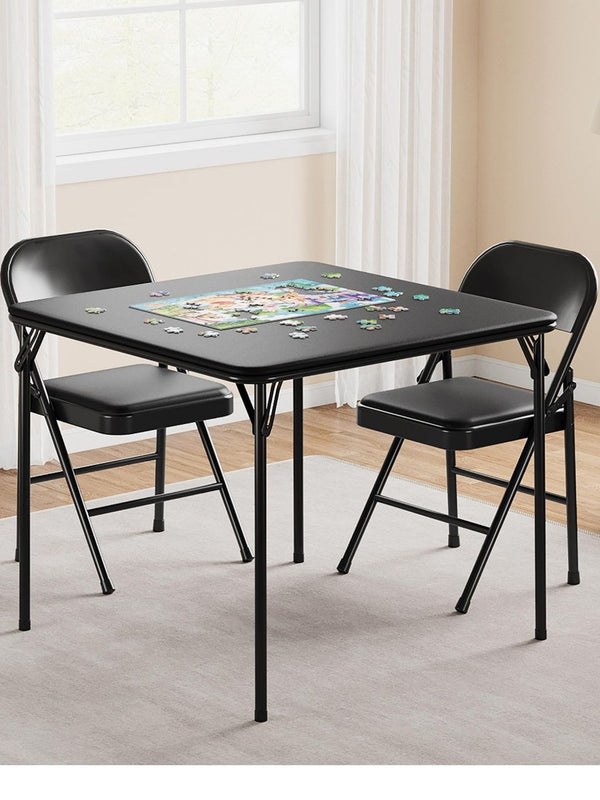 A black square folding table with two matching black folding chairs in a bright room with white curtains, wood floors, and a rug. A colorful jigsaw puzzle is spread on the tabletop.