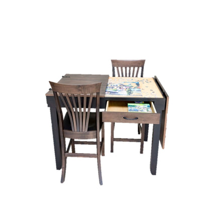 A dark wood jigsaw puzzle table with two matching wooden chairs, featuring a recessed jigsaw puzzle playing area and an open drawer holding jigsaw puzzle pieces with a plain white background.