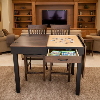 A dark wood and light wood jigsaw puzzle table with two chairs tucked underneath, shown in a living room setting with a TV and bookshelves in the background. One drawer is open, revealing a jigsaw puzzle boxes inside with jigsaw puzzle pieces on the open side of the tabletop.
