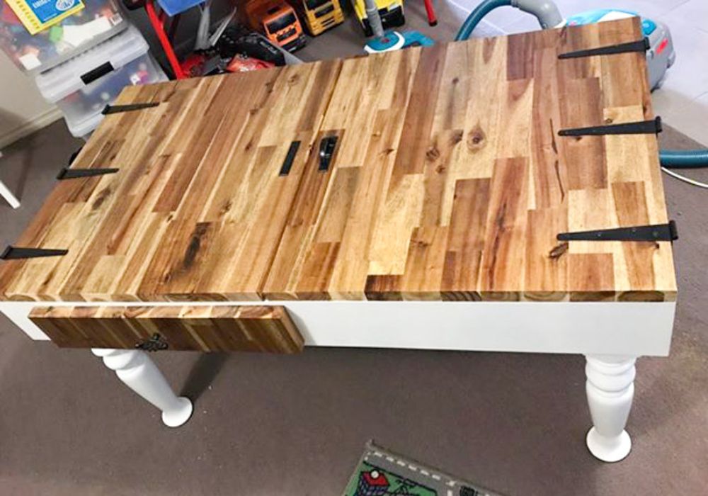 A rustic wooden jigsaw puzzle table with a multi-toned natural wood top, black metal brackets, a matching wood drawer front, and white turned legs, photographed in a playroom environment.
