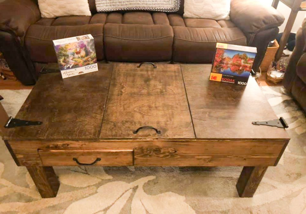 A rustic wooden jigsaw puzzle coffee table with a lift-top design, shown in a living room setting with a brown leather sofa and patterned rug behind it. Two jigsaw puzzle boxes are placed on the table, one on each side.