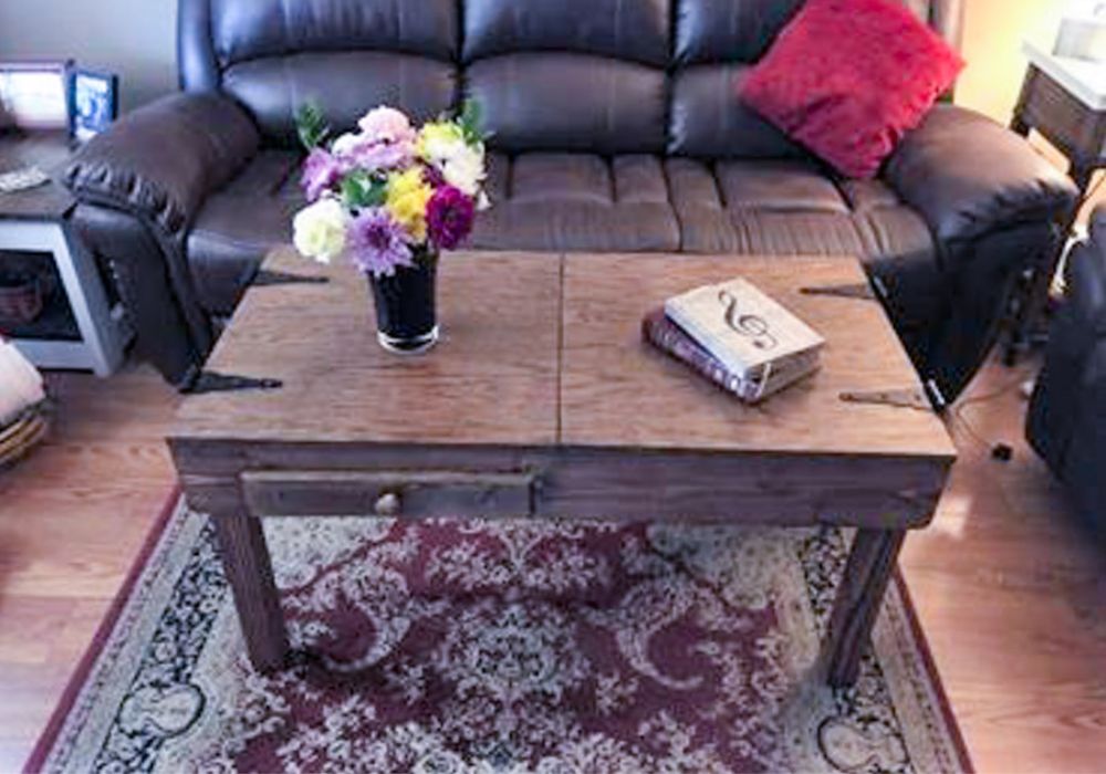 A rustic wooden jigsaw puzzle coffee table with a single drawer and metal corner brackets, placed in a living room on a patterned rug in front of a dark leather sofa.
