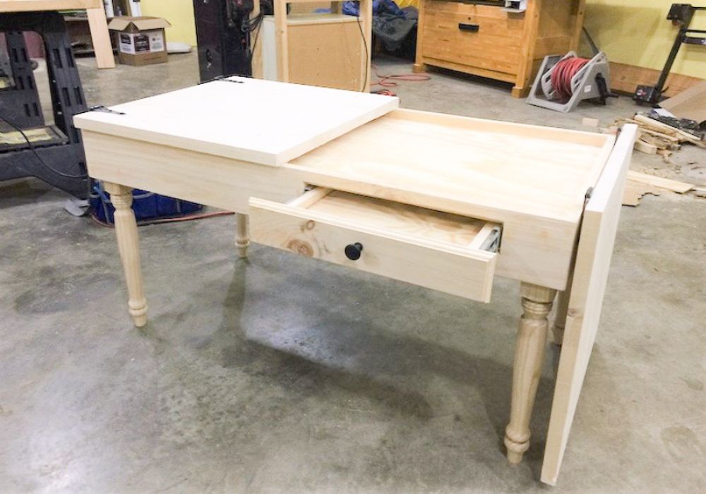 A light natural wood jigsaw puzzle table with a lift-top section and an open drawer, photographed in a workshop environment.