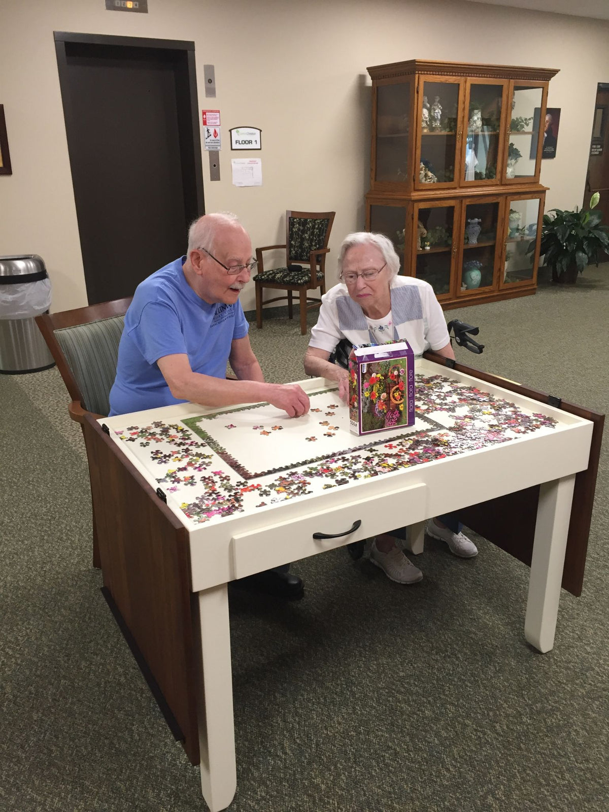 An elderly man and woman sitting at a white wood jigsaw puzzle table, working on a jigsaw puzzle together in a community center or senior center room.
