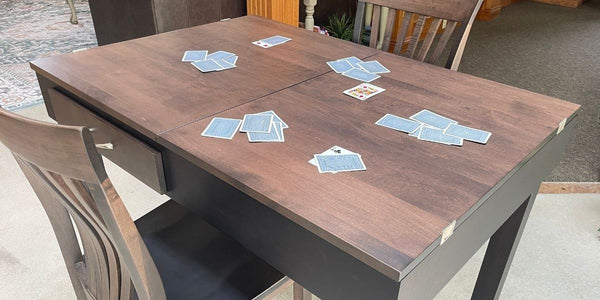 A dark brown wooden jigsaw puzzle table with a dark gray base, surrounded by wooden dining chairs. Several playing cards are scattered on the tabletop.