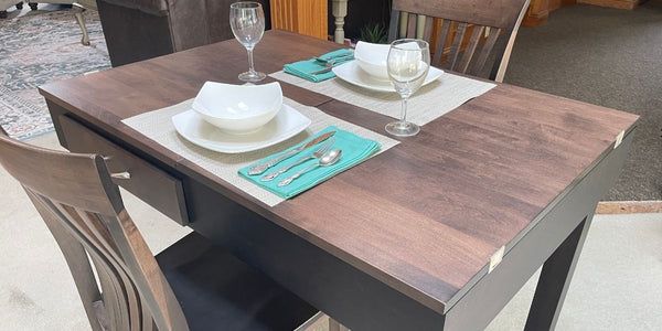 A rectangular wooden jigsaw puzzle table with a dark brown wood-grain top and dark gray legs, accompanied by matching wooden chairs. The table is set with white plates, white bowls, teal napkins, silver cutlery, and wine glasses.