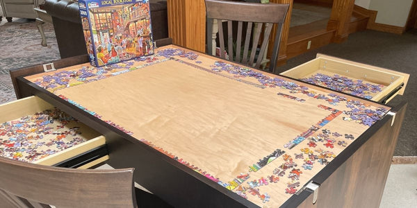 A wooden puzzle table with multiple drawers opened to reveal filled jigsaw puzzle compartments, with a puzzle box standing at the top left. The photo is taken in a home interior with chairs and furniture visible around the table.