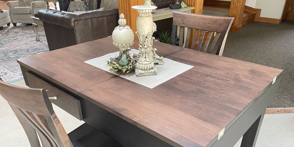 A dark brown wooden jigsaw puzzle table with a dark gray base, shown with wooden chairs in a furniture showroom setting. The table is styled with a white placemat and two decorative cream-colored vases.