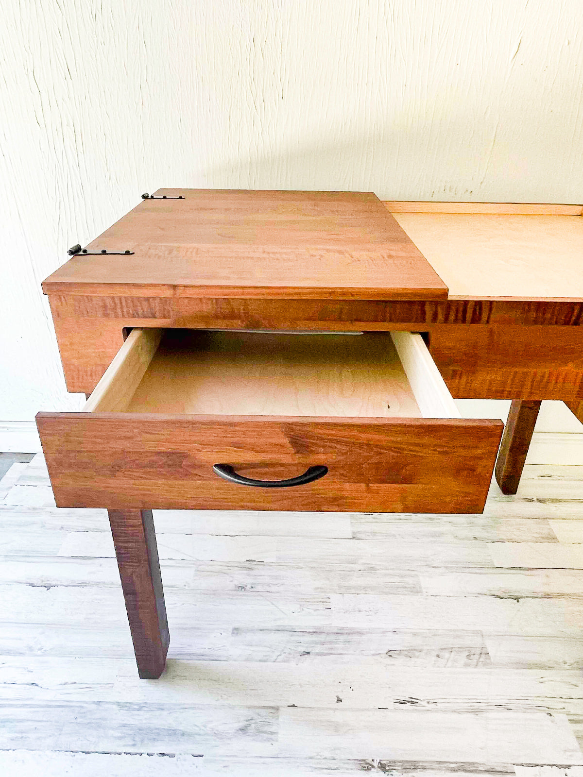 A wooden jigsaw puzzle table  with a warm medium-brown finish and a light natural wood drawer interior, shown with one drawer pulled open. The desk has a dark metal pull handle and is photographed against a light textured wall and whitewashed wood floor.