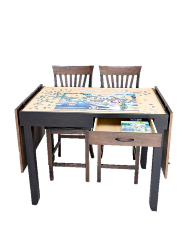 A semi custom jigsaw puzzle table with two matching chairs, featuring a light wood tabletop with a colorful jigsaw puzzle, a pull-out drawer, and fold-open tabletop, shown on a plain white background.