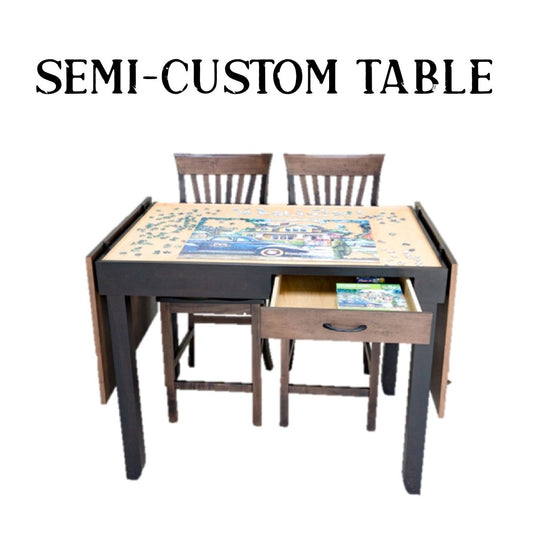 A semi-custom wooden jigsaw puzzle table set with two matching chairs, featuring a dark frame, light wood top, and an open drawer. The table is shown with a colorful puzzle spread across the surface.