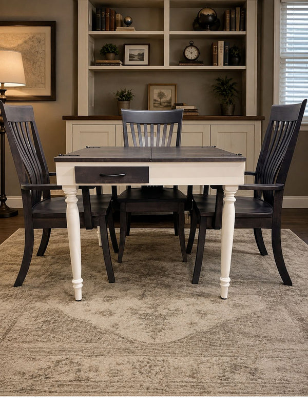A square jigsaw puzzle table with a white and dark gray painted finish, featuring a single drawer on the front, surrounded by three dark gray spindle-back chairs. The set is styled on a patterned beige rug in a dining room with a built-in bookshelf in the background.