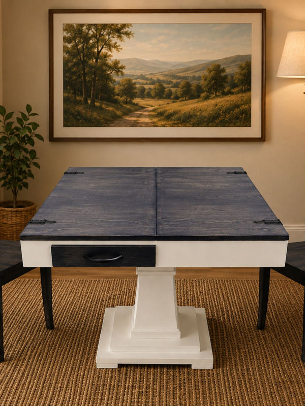 A square jigsaw puzzle table with a dark gray stained top and a white pedestal base, shown in a styled room setting with a framed landscape painting on the wall, a plant, and a lamp.